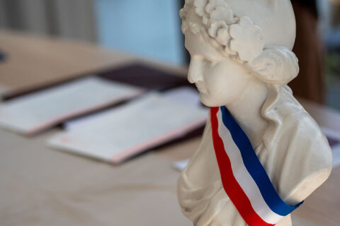 Buste de Marianne portant l'écharpe tricolore avec un parapheur en arrière plan évoquant une réunion du conseil municipal