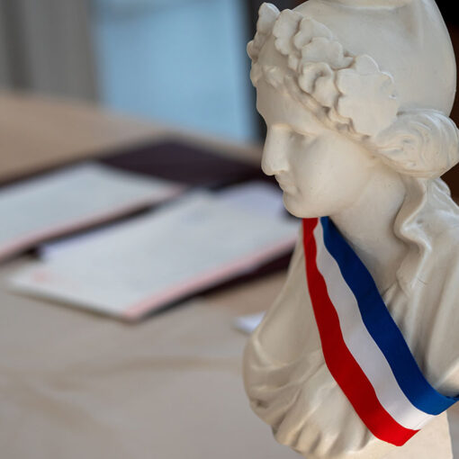 Buste de Marianne portant l'écharpe tricolore avec un parapheur en arrière plan évoquant une réunion du conseil municipal